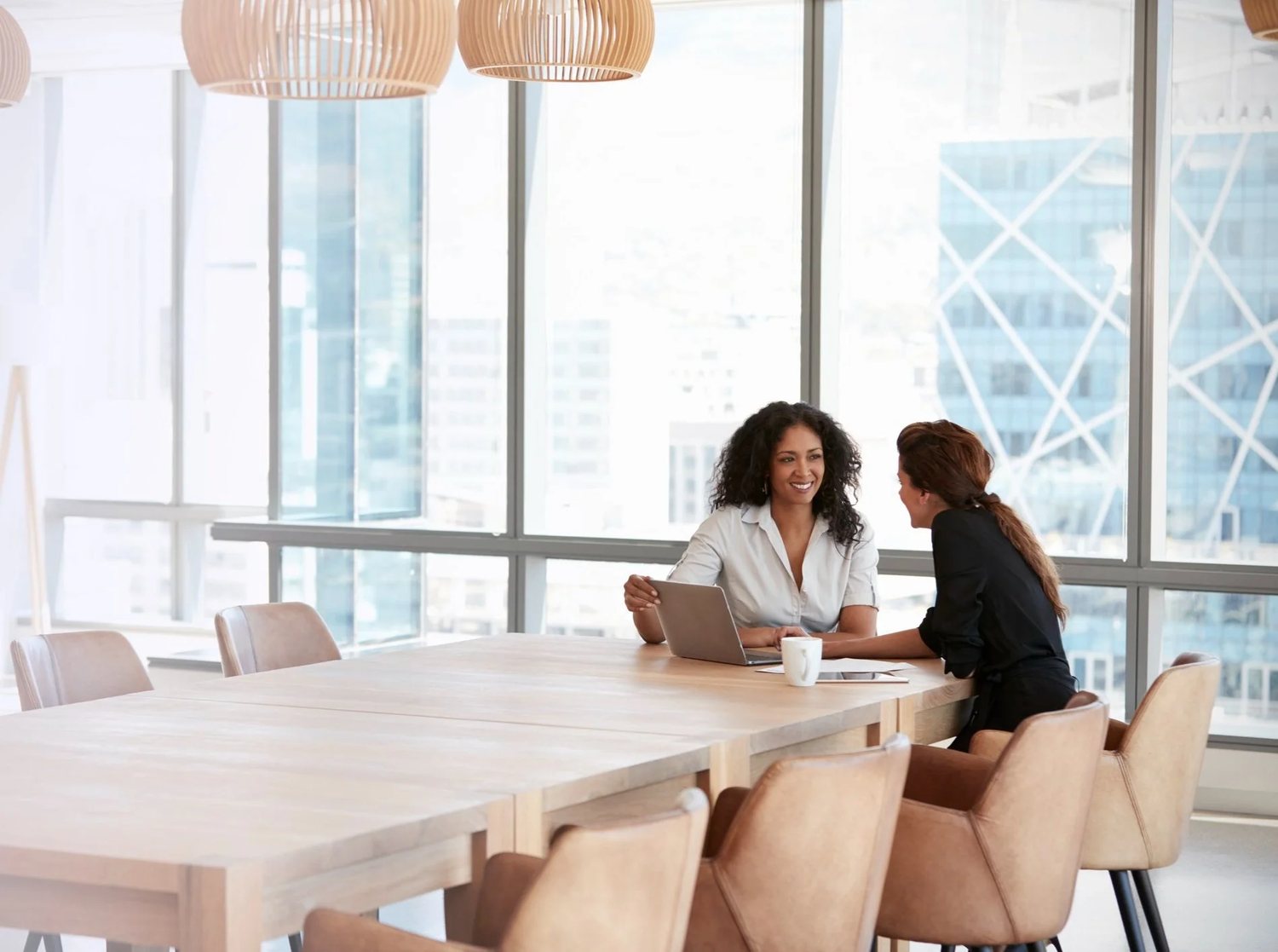 Two people working together at a meeting table beside a wall of windows.